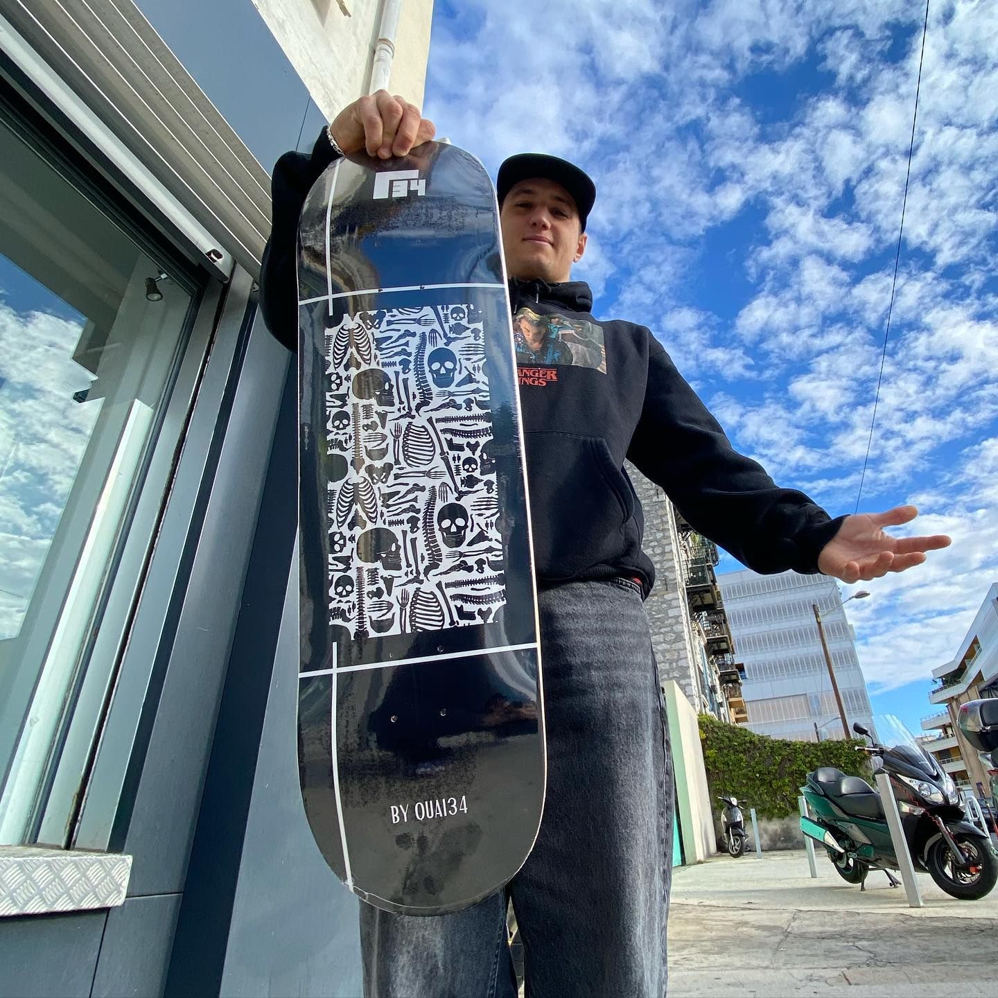 Un homme tenant un skateboard noir avec un motif blanc, à l'extérieur près d'un bâtiment, par une journée ensoleillée.