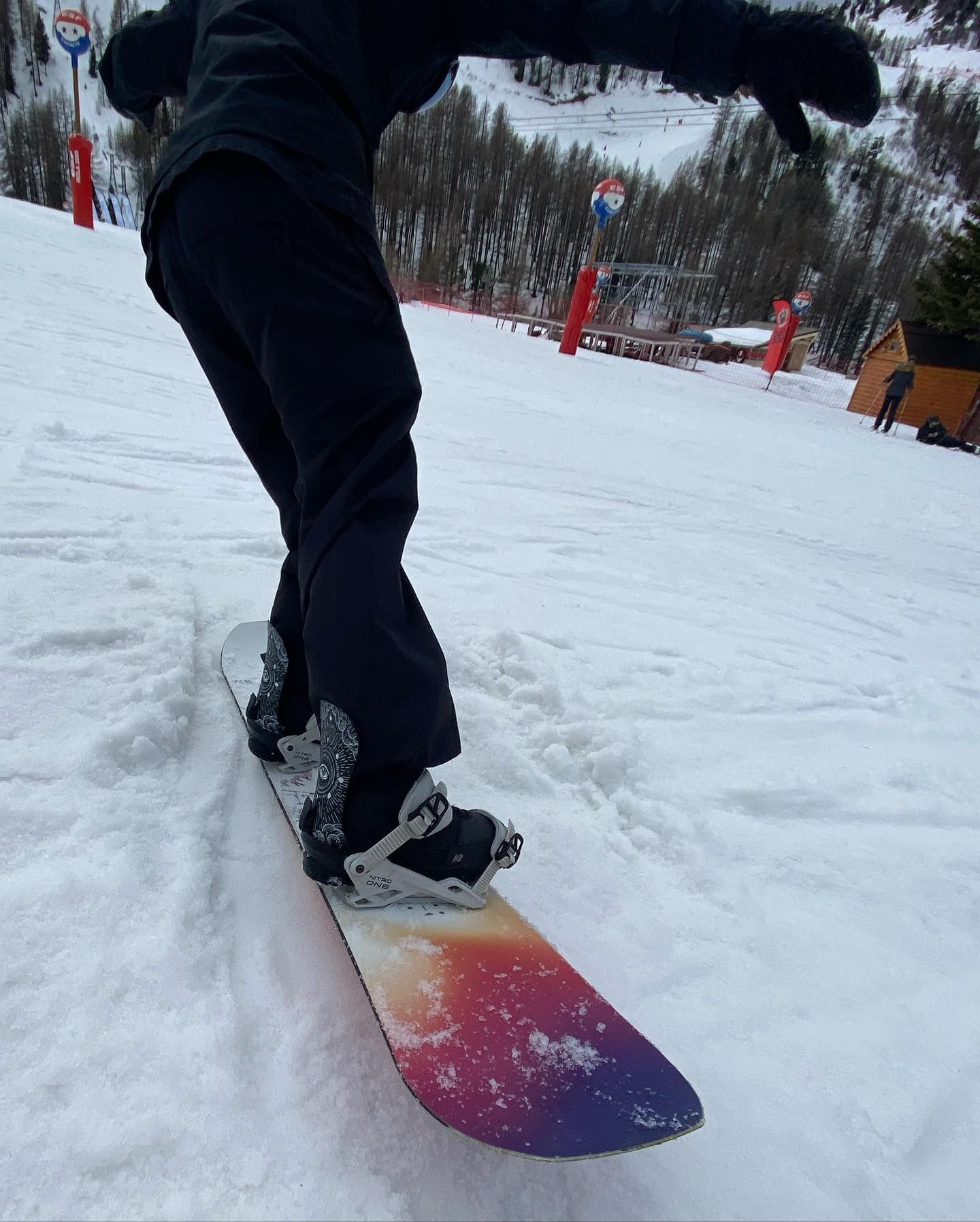 Un snowboardeur dévale une pente enneigée, vêtu d'un pantalon et d'une veste foncés. Sa planche présente un dégradé de couleurs.