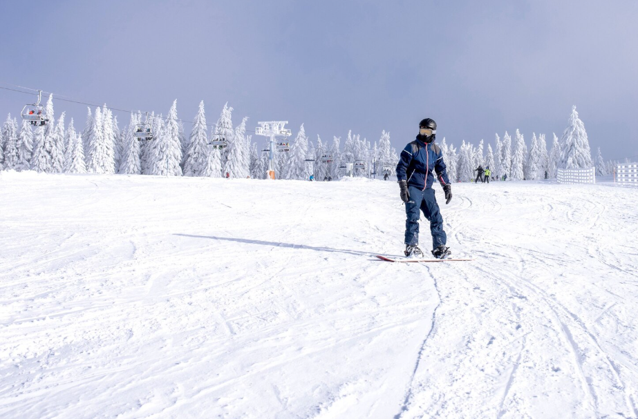 Personne en snowboard sur une pente enneigée, arbres et télésiège en arrière-plan, ciel couvert.