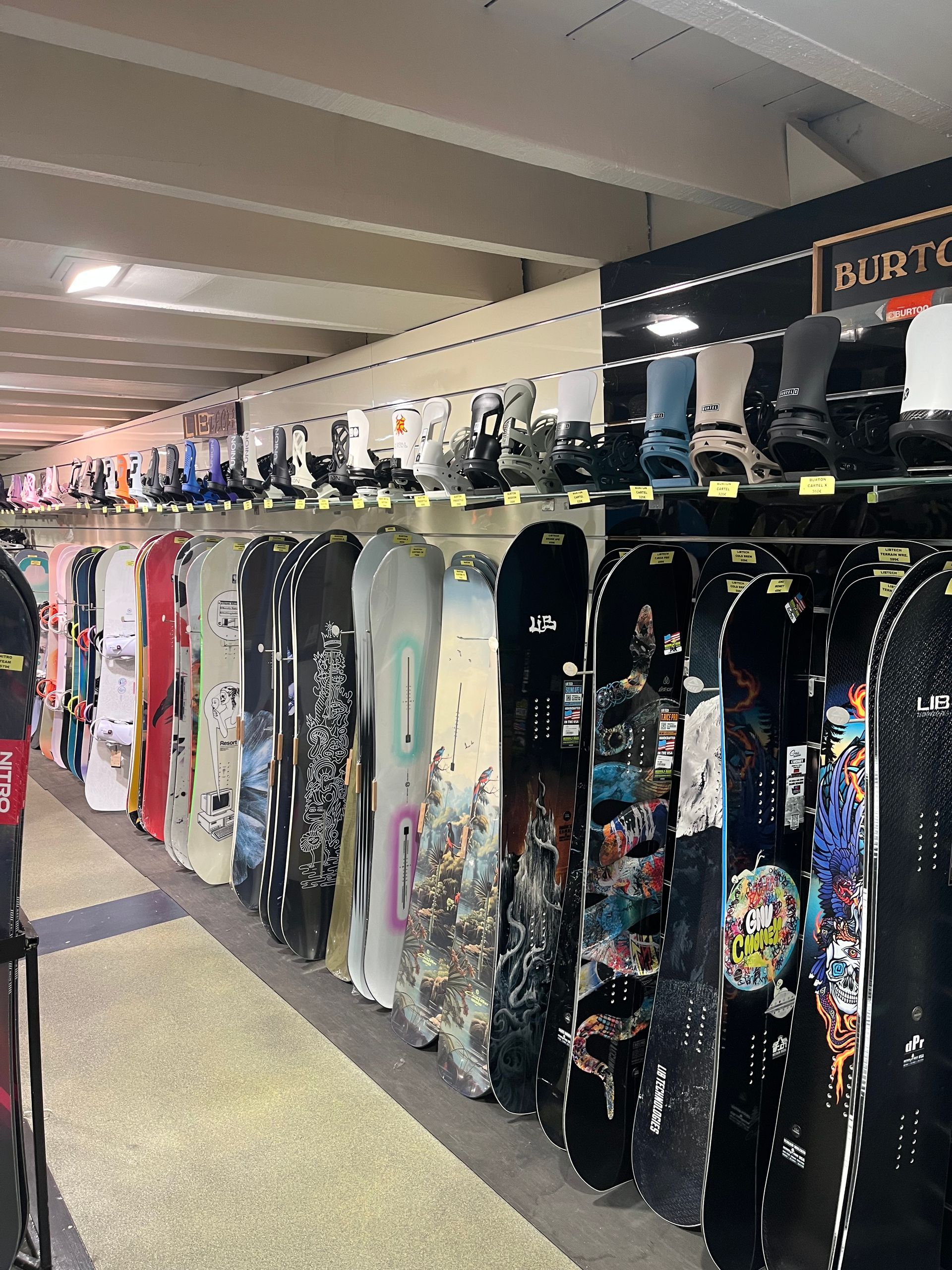 Des rangées de snowboards sont exposées dans un magasin. Des couleurs et des modèles variés tapissent les murs, avec des étagères au-dessus desquelles sont présentés les modèles de bottes.