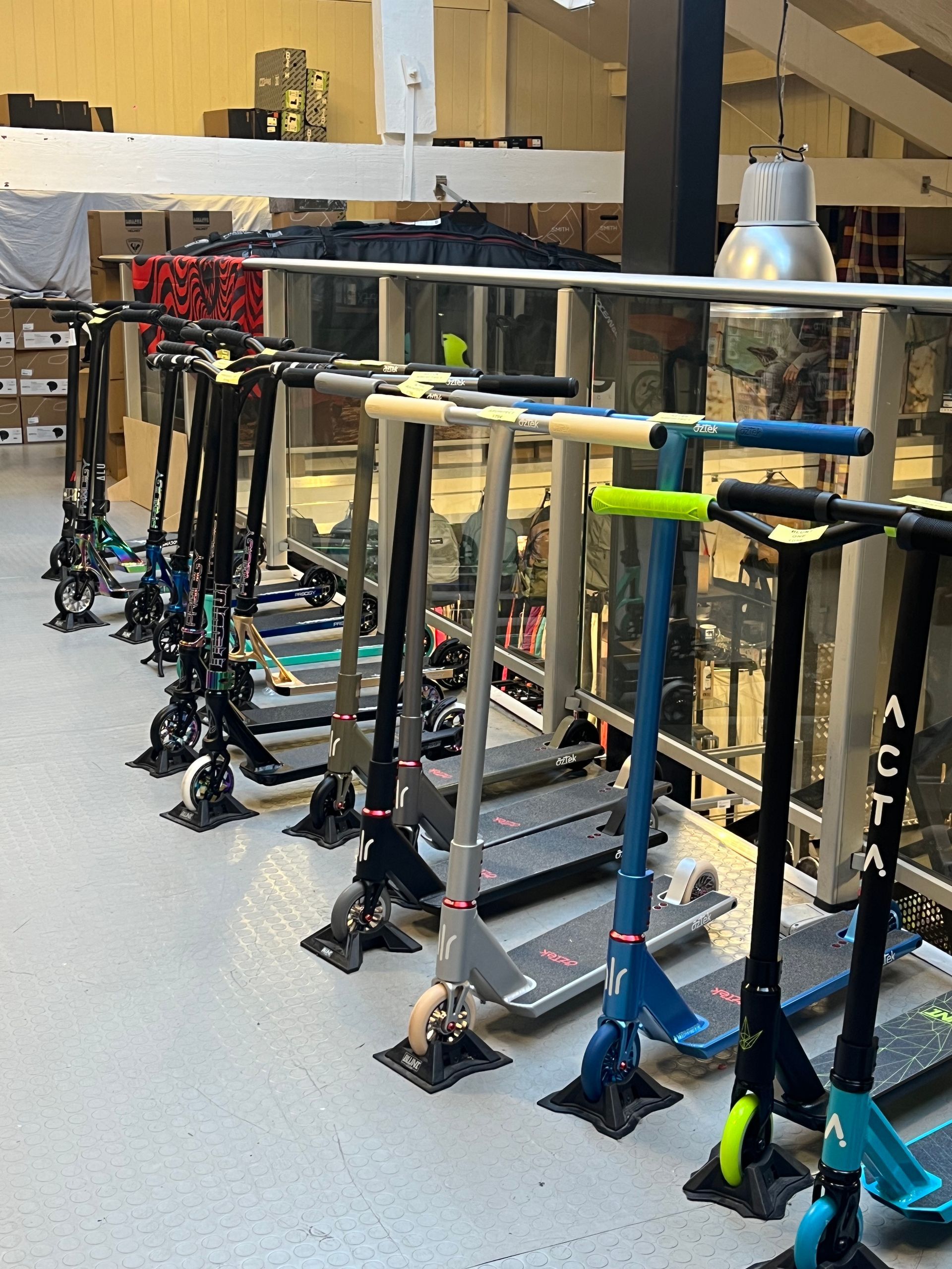 Une rangée de scooters est exposée à l'intérieur d'un magasin. De couleurs et de styles variés, près d'une rambarde et d'étagères.