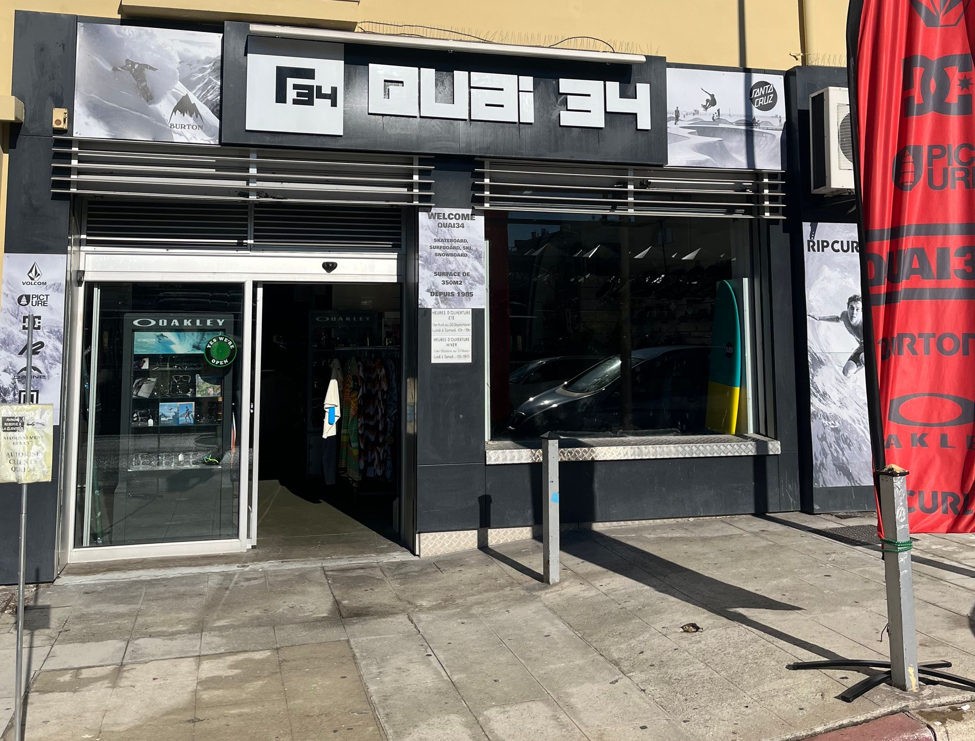 Façade de magasin avec enseigne « Quai 34 ». Extérieur noir, vitrines, porte ouverte et banderole rouge.