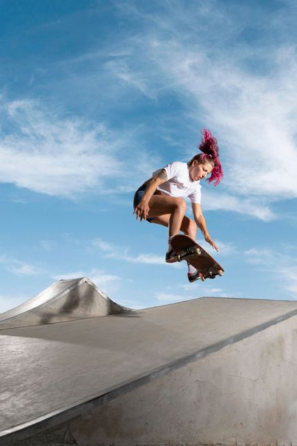 Une personne en skateboard saute par-dessus une rampe en béton sur fond de ciel bleu. Cheveux roses.
