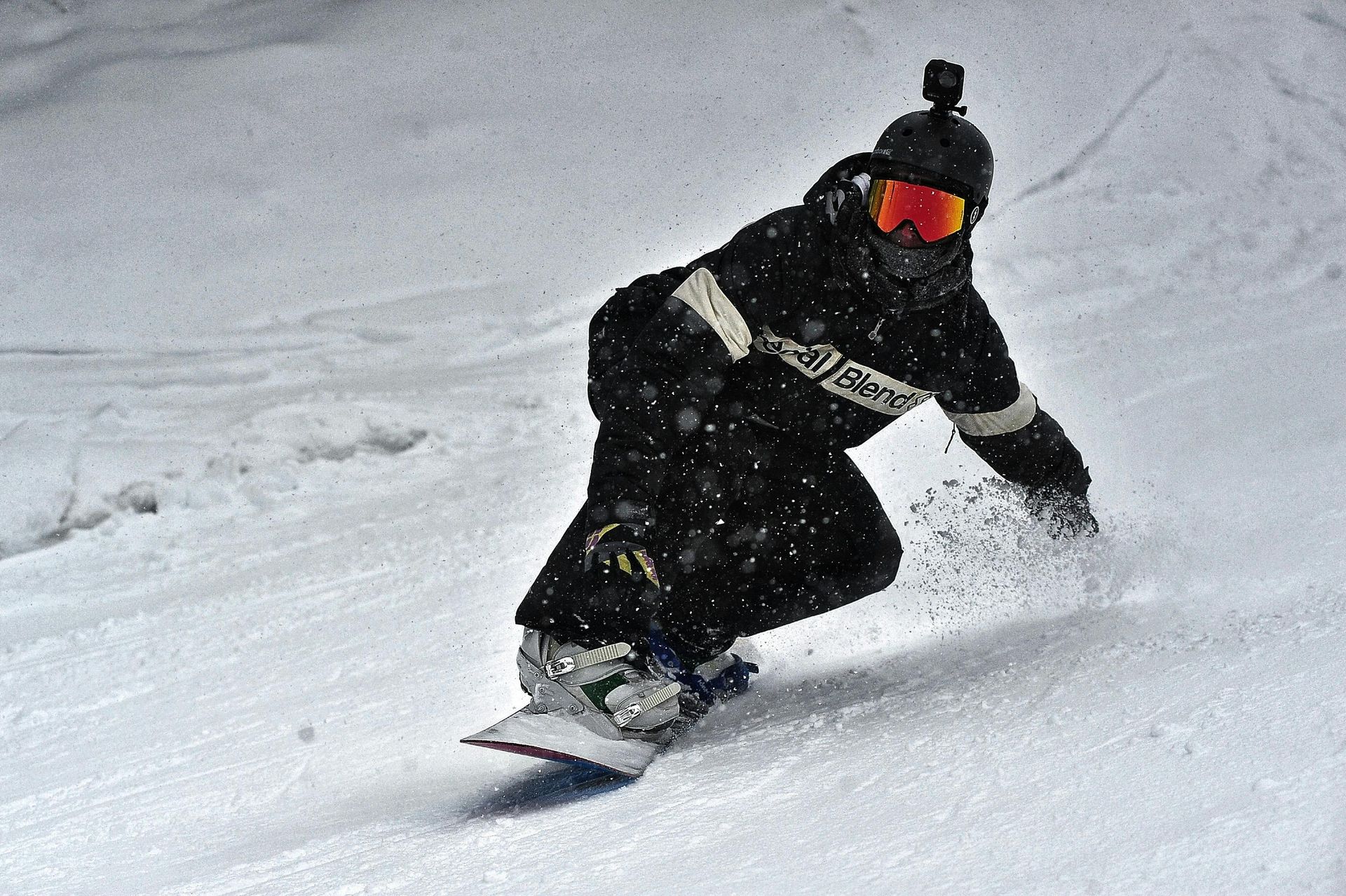 Un snowboardeur dévale une pente enneigée, vêtu d'une veste noire, de lunettes de ski orange et d'un casque équipé d'une caméra.