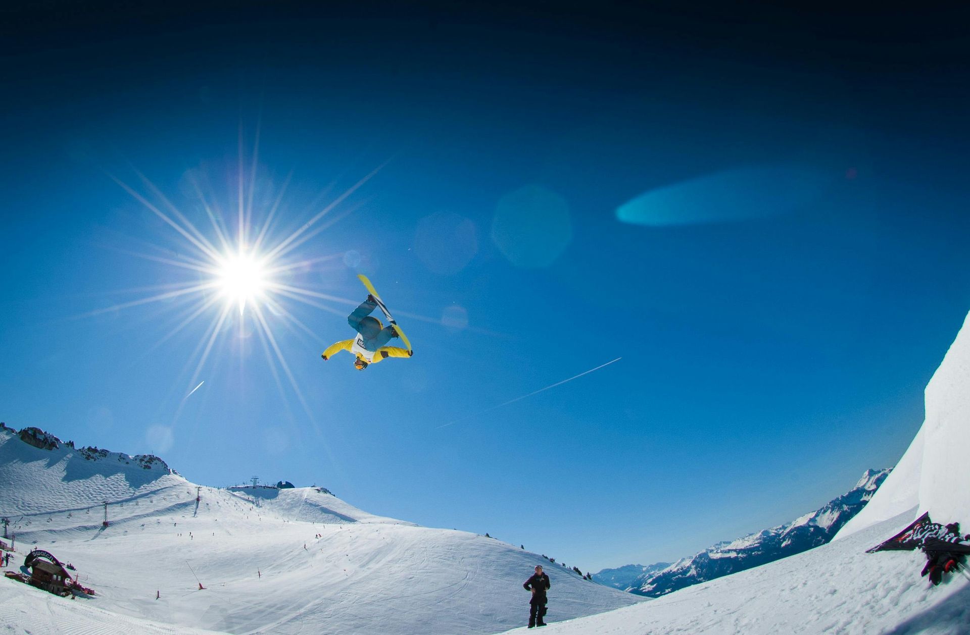 Snowboardeur en plein vol, à contre-jour, au-dessus d'une chaîne de montagnes enneigées.