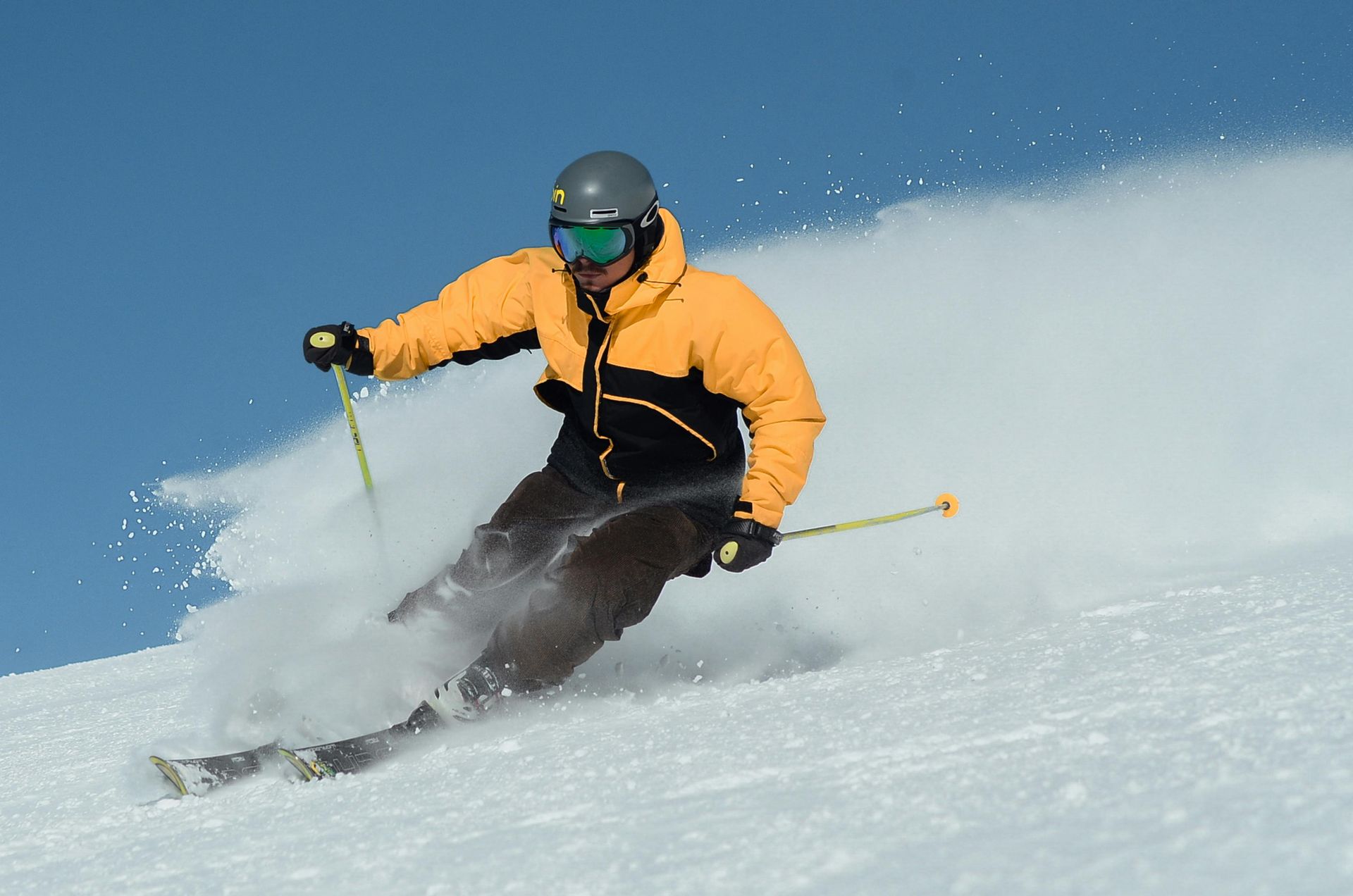 Skieur en veste jaune dévalant une pente enneigée, soulevant un nuage de neige, journée ensoleillée.