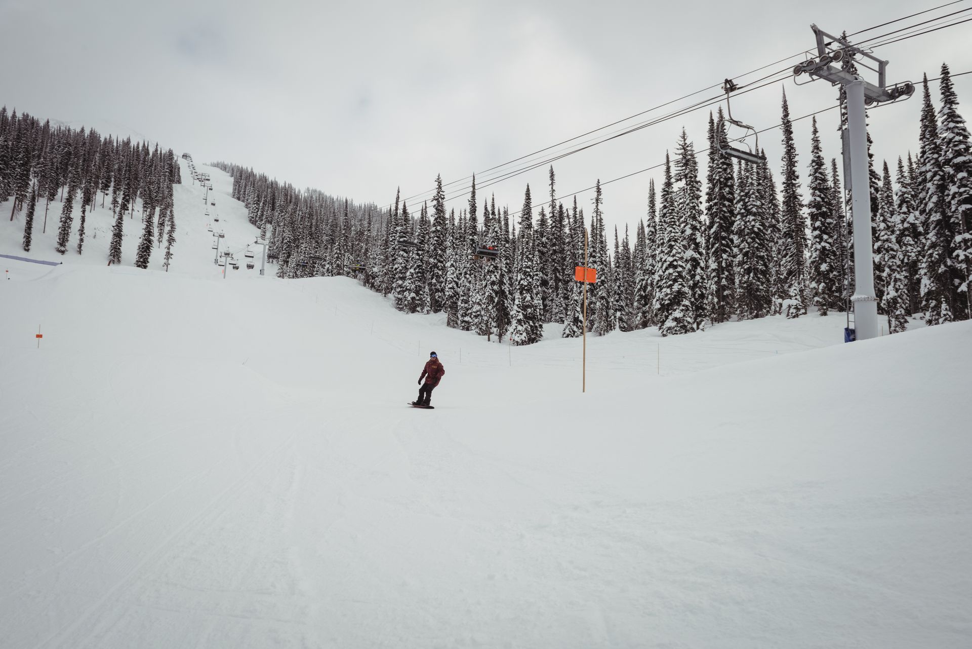Un snowboardeur sur une pente enneigée près d'un télésiège et de conifères sous un ciel nuageux.