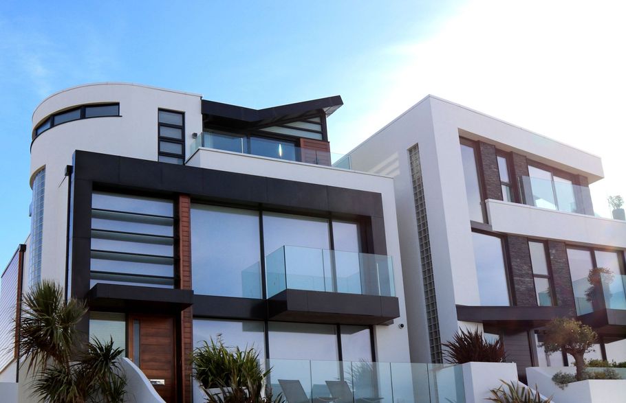 Casa de lujo moderna en blanco y gris con grandes ventanales y un balcón bajo el cielo azul.