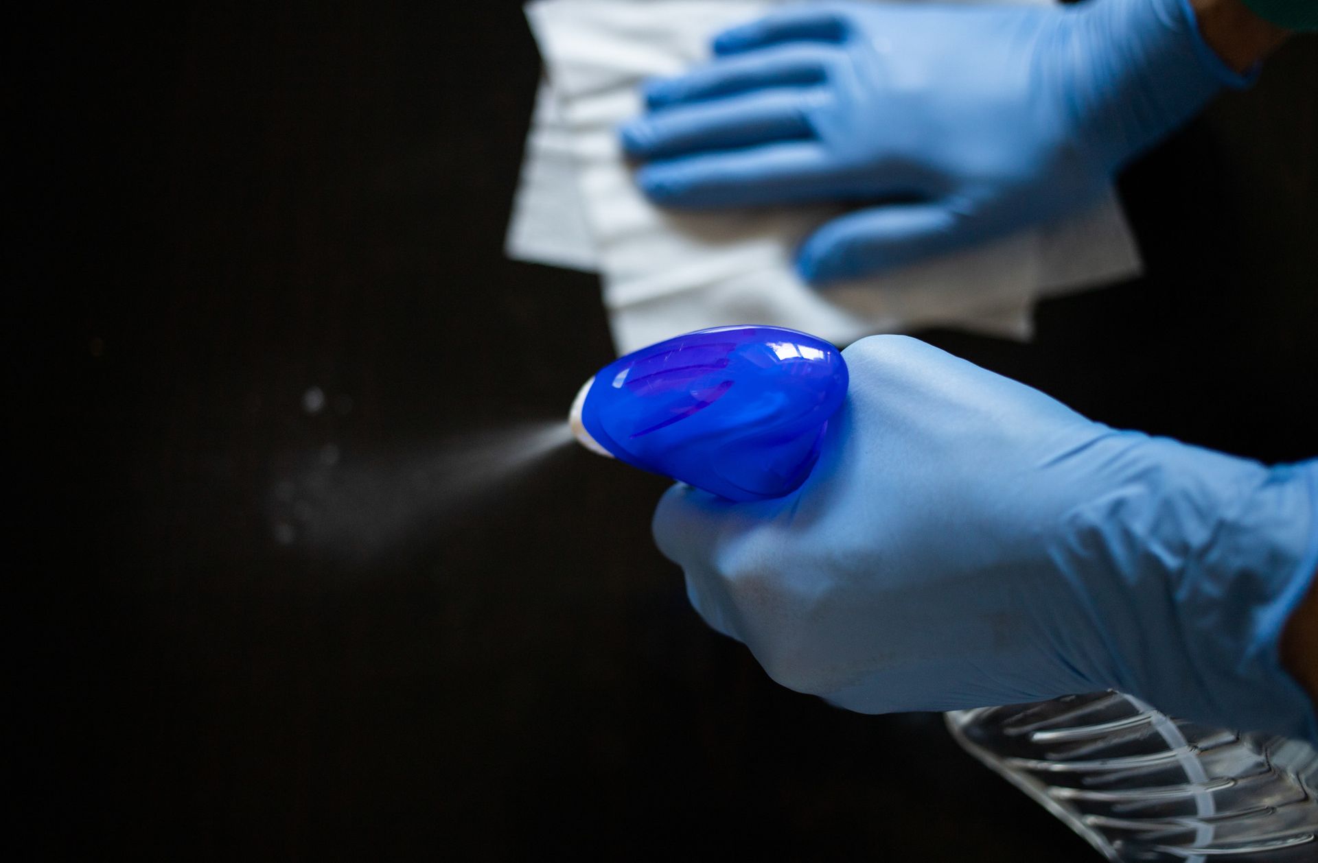 Manos con guantes azules rociando desinfectante y limpiando la superficie con un paño.