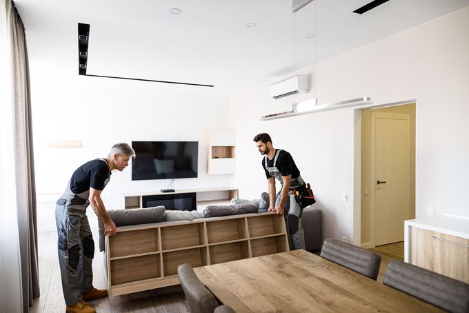 Dos trabajadores levantan un sofá seccional en una sala de estar luminosa con TV y mesa de comedor.