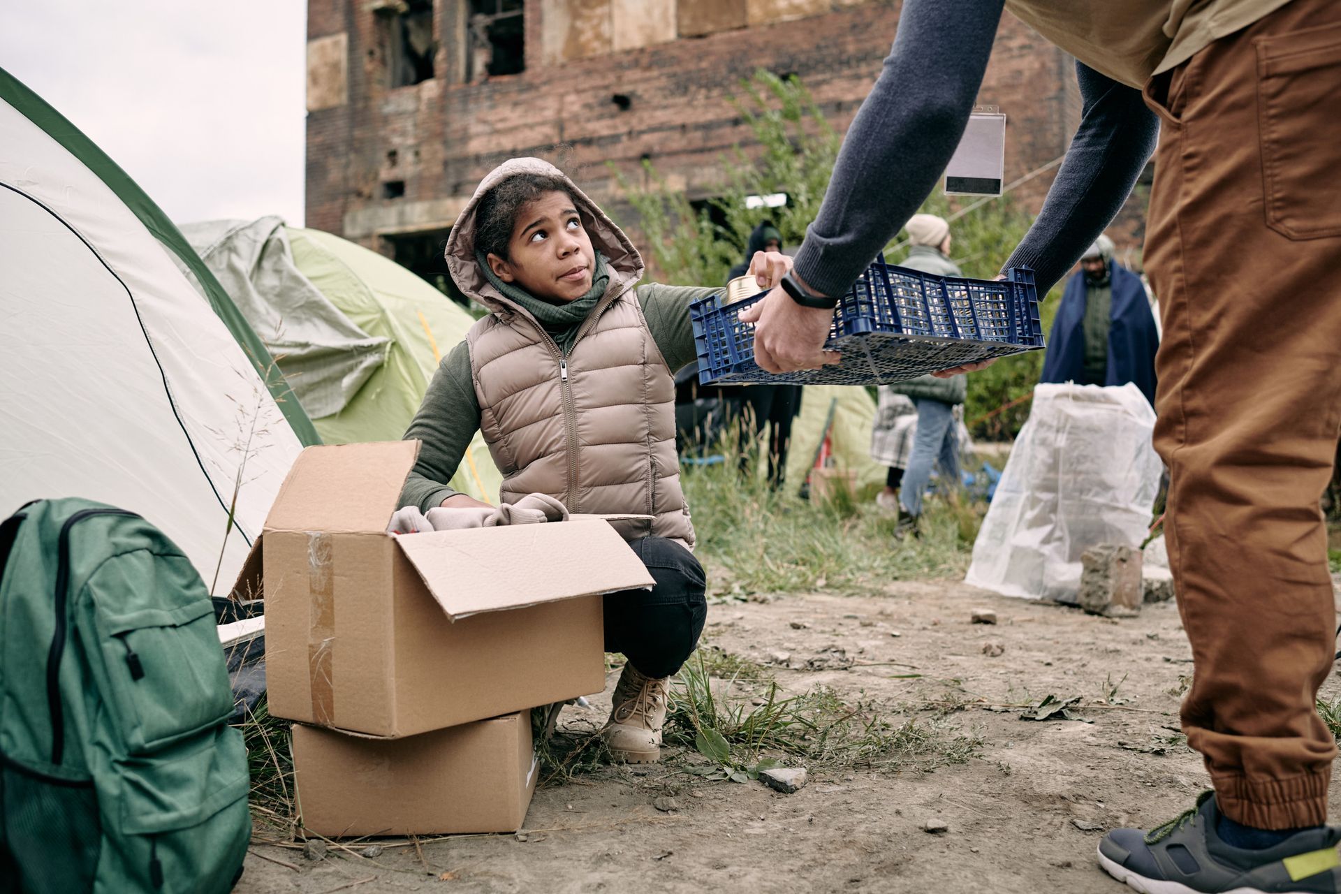 Persona entregando una canasta a un niño cerca de una tienda de campaña y cajas en un área de edificio dañado.