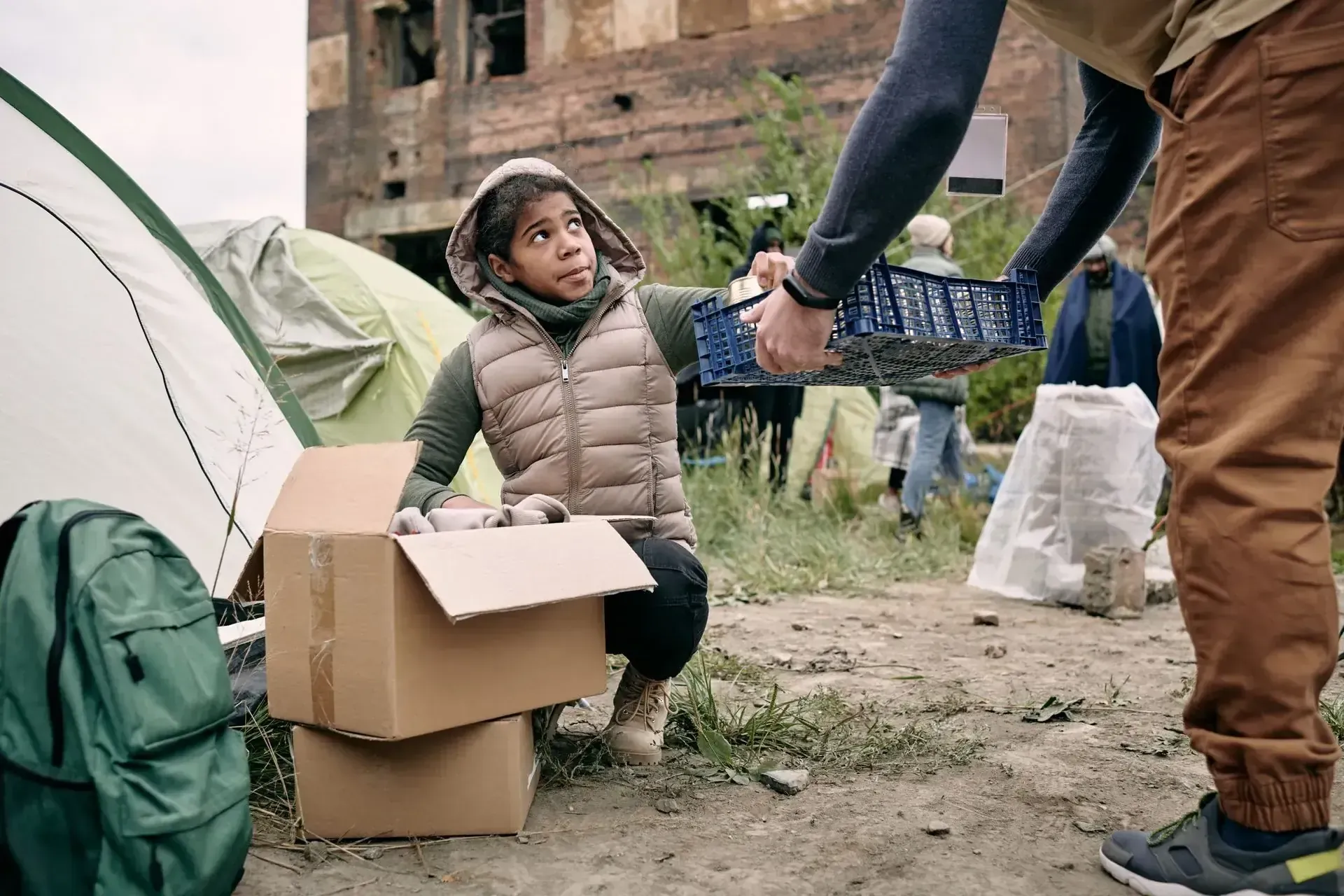 Persona entregando una canasta a un niño cerca de una tienda de campaña y cajas en un área de edificio dañado.
