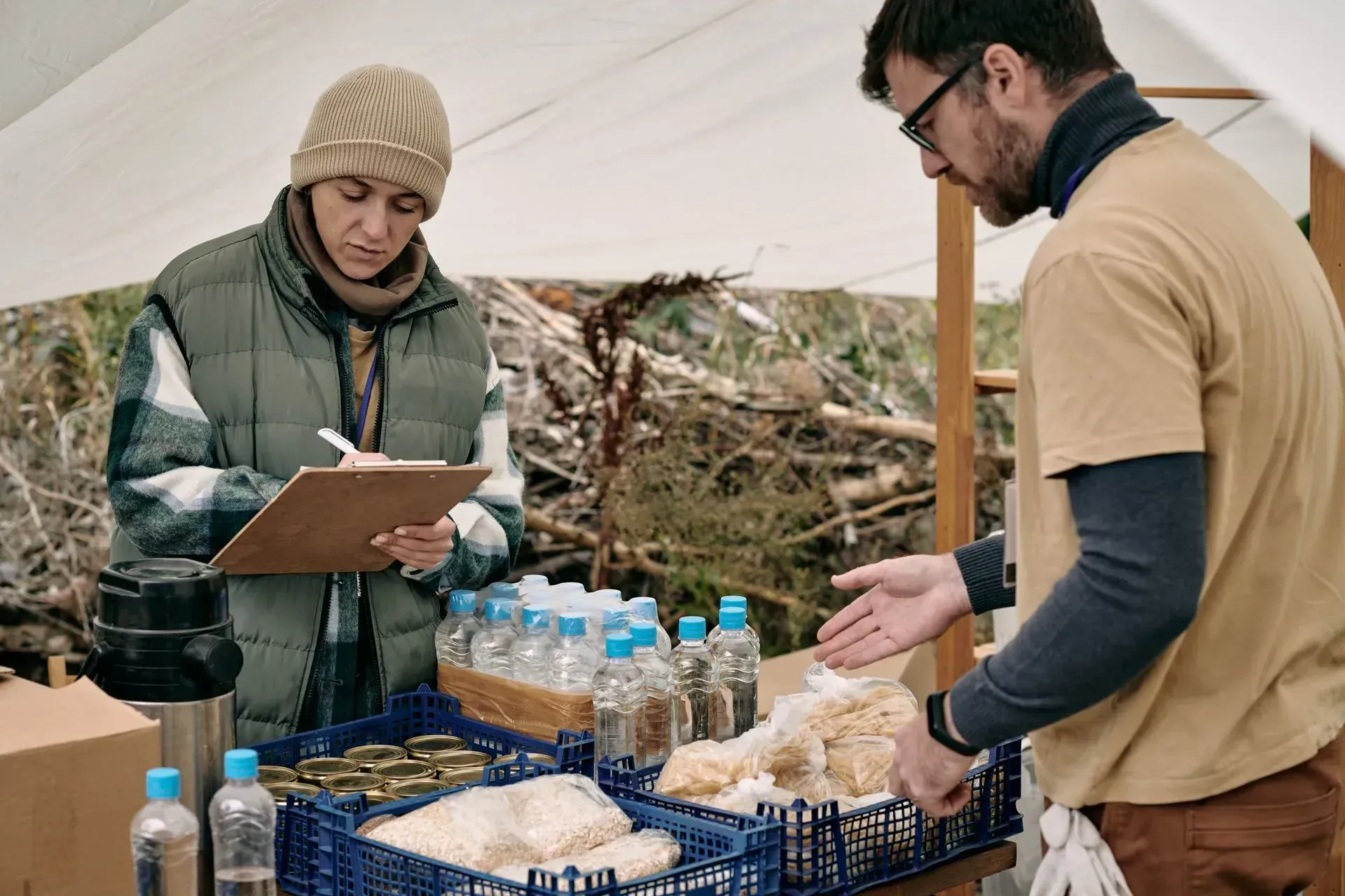 Mujer con portapapeles y hombre en una mesa, posiblemente en un sitio de distribución de alimentos, bajo un dosel blanco.