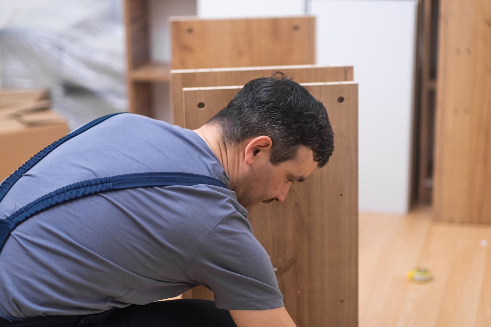 Hombre ensamblando muebles de madera en un interior. Está concentrado, con tirantes azules, cerca de tablas de color marrón claro.