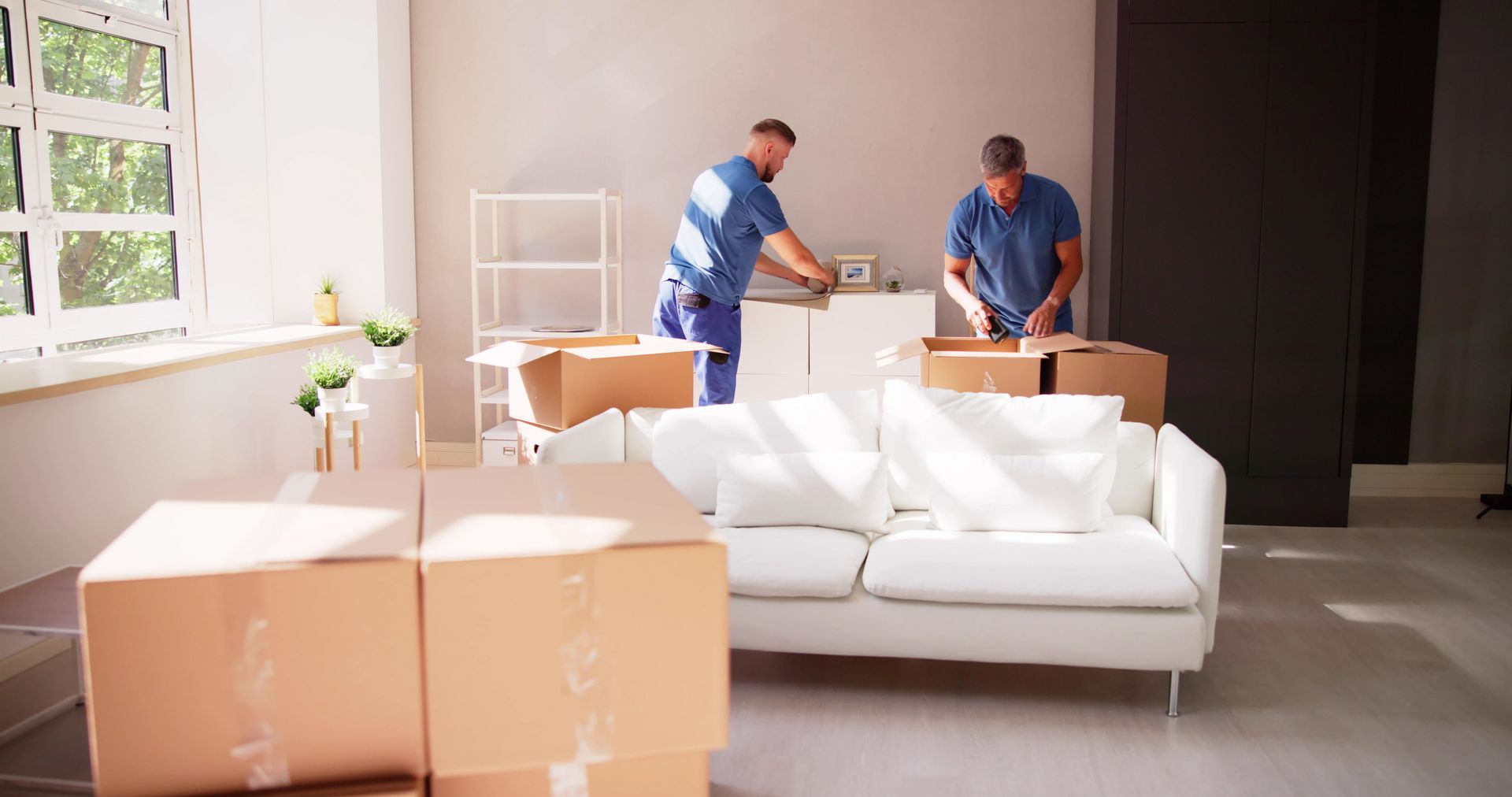 Dos personas empacando cajas en una habitación bien iluminada con un sofá blanco y una ventana grande.