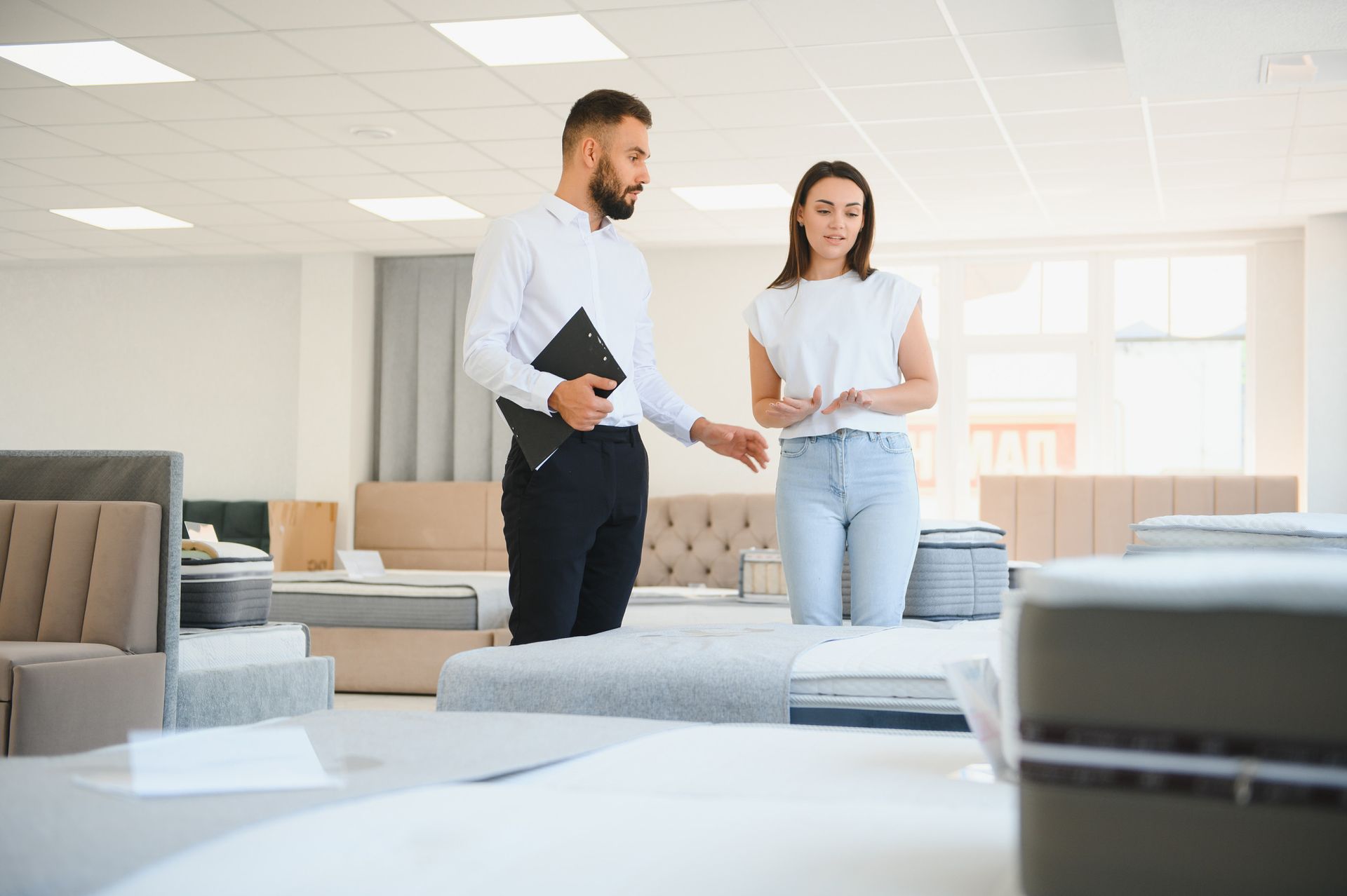 Hombre con camisa blanca mostrando un colchón a una mujer con jeans de color claro en una tienda de muebles.