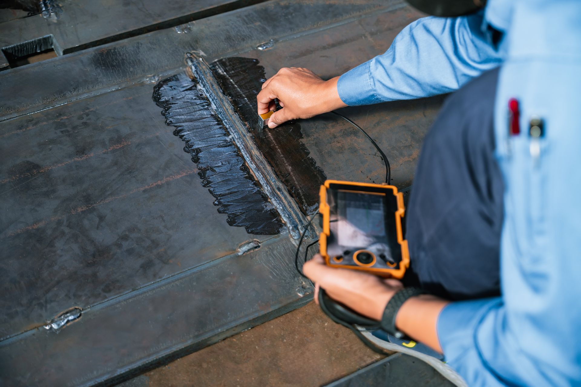 Un ouvrier inspecte une surface métallique à l'aide d'un appareil portatif et applique du mastic le long d'un joint.