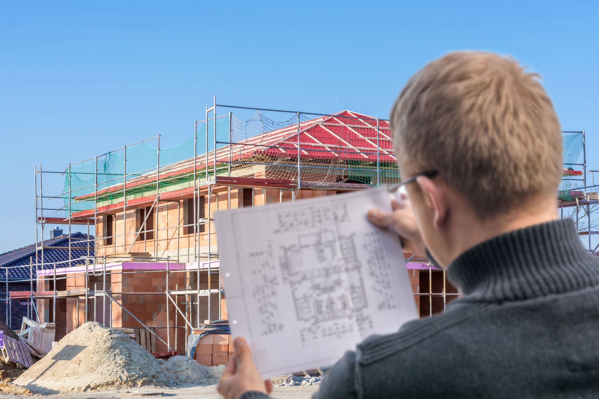 Un ouvrier du bâtiment examine les plans d'une maison sur un chantier où se trouvent des échafaudages et une toiture partiellement construite.