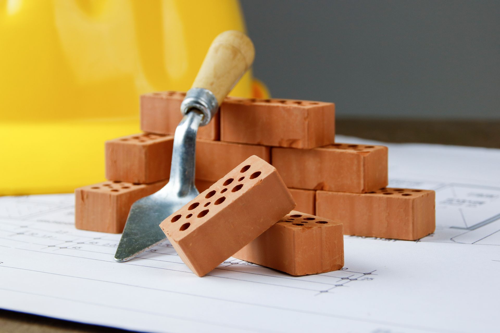 Truelle de maçon à côté de briques rouges empilées sur un plan, avec un casque de chantier jaune en arrière-plan.