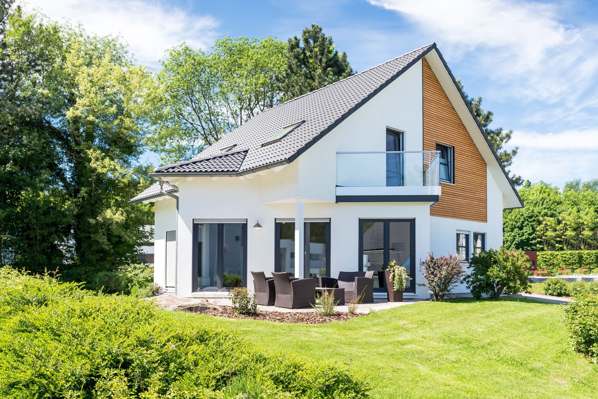 Maison blanche moderne avec toit foncé, terrasse et pelouse verte baignée de soleil.