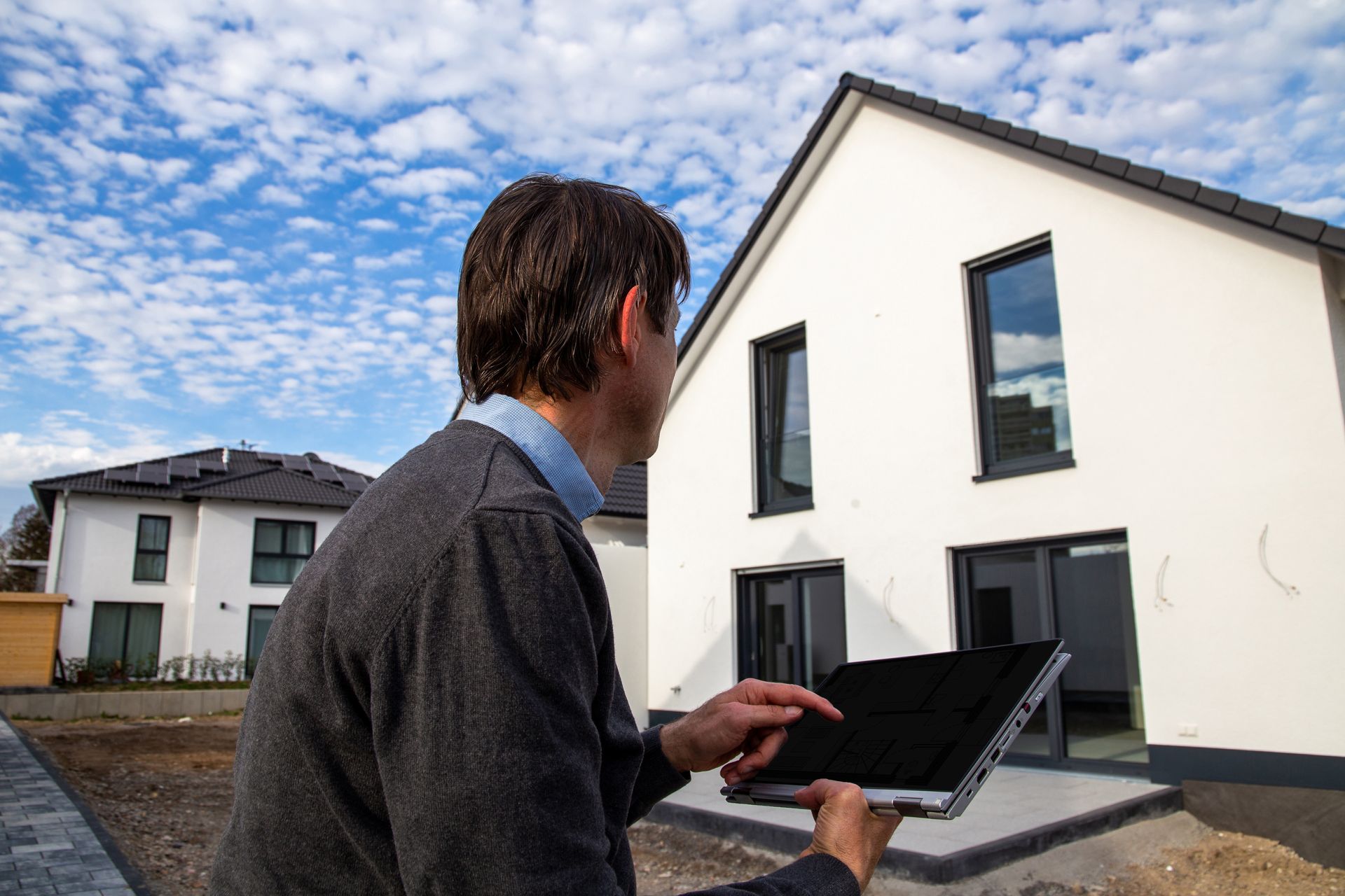 Une personne tenant un ordinateur portable et regardant l'extérieur d'une maison moderne dans un quartier résidentiel