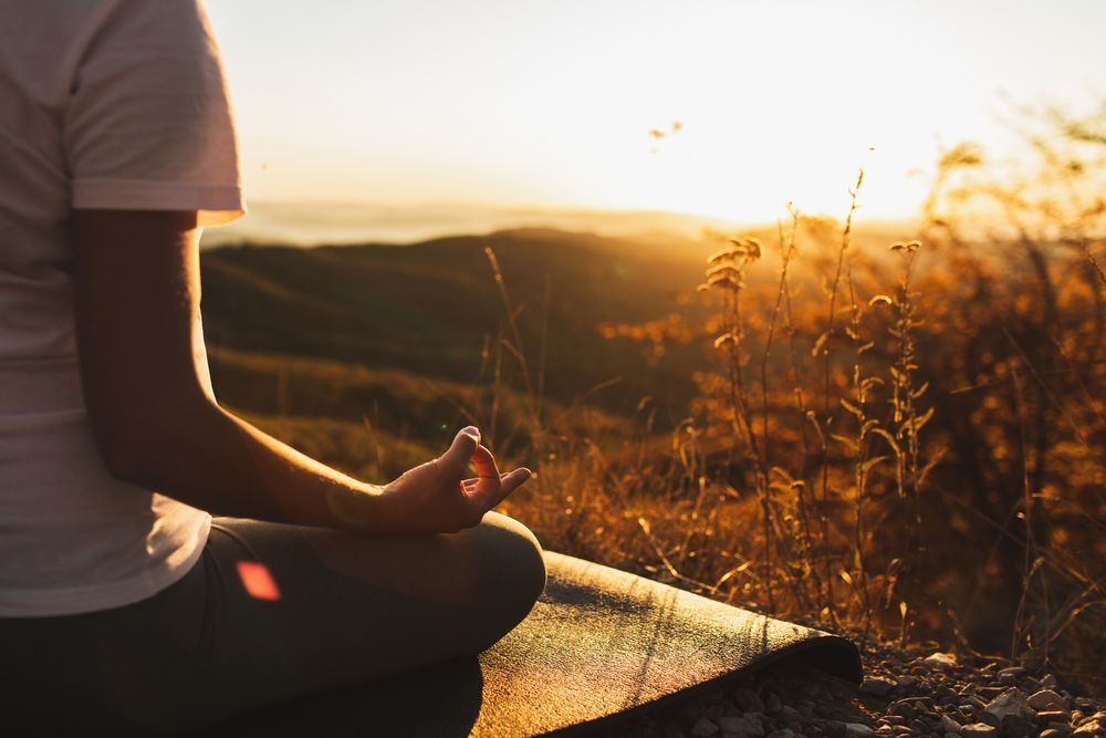 Persona meditando al aire libre durante la puesta de sol, con las manos en posición mudra, luz solar naranja.