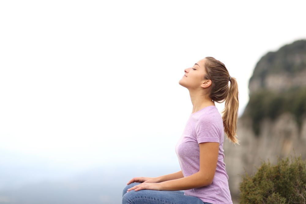 Una mujer con una camisa morada está sentada con los ojos cerrados, respirando profundamente, en la cima de una montaña con un cielo nublado de fondo.