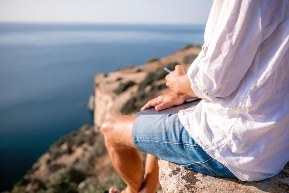 Una persona está sentada al borde de un acantilado, mirando su portátil mientras contempla el océano azul. Lleva una camisa blanca y pantalones cortos vaqueros.