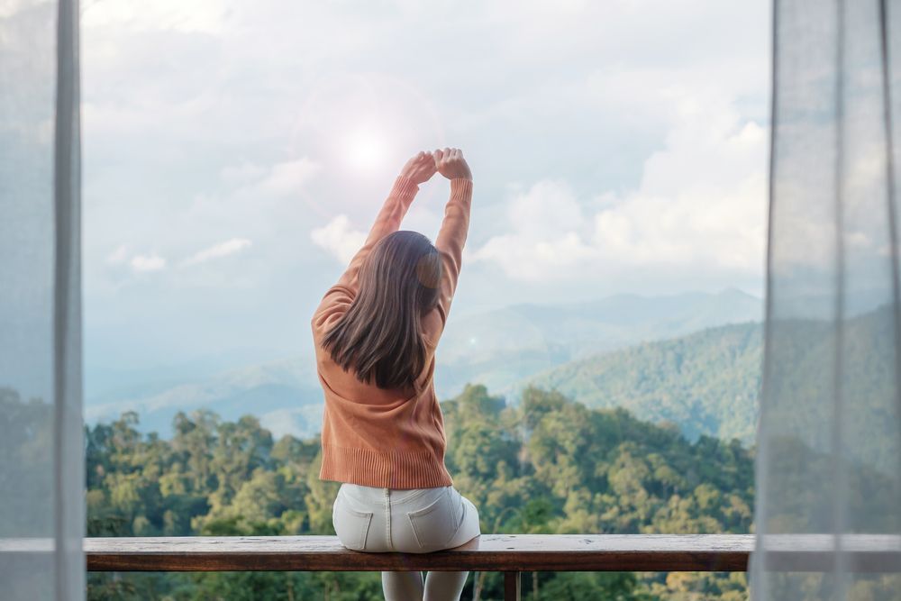 Mujer estirándose en un balcón con vistas a un paisaje montañoso, bañado por la luz del sol.