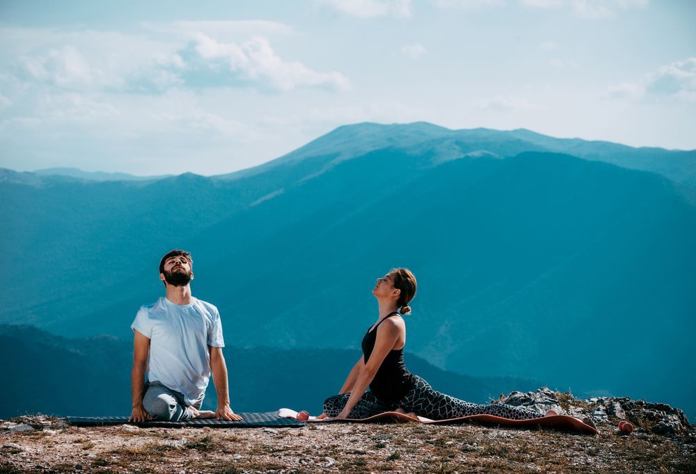 Dos personas practicando yoga al aire libre con montañas de fondo. El hombre y la mujer se estiran sobre una esterilla y una manta.