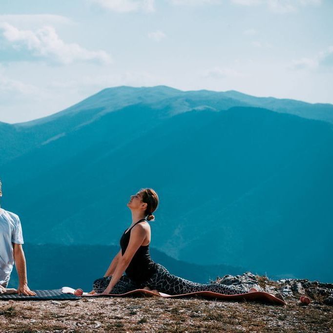 Dos personas haciendo yoga en la cima de una montaña, una en posición de estocada y la otra fuera de cámara, con un fondo de montañas azules y cielo.