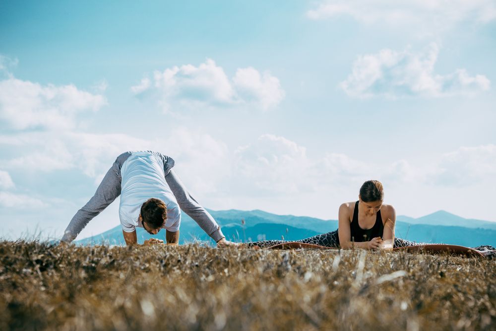 Dos personas practicando yoga al aire libre: una en posición de flexión hacia adelante con las piernas abiertas, la otra en decúbito prono con los brazos extendidos. Montañas y cielo azul de fondo.