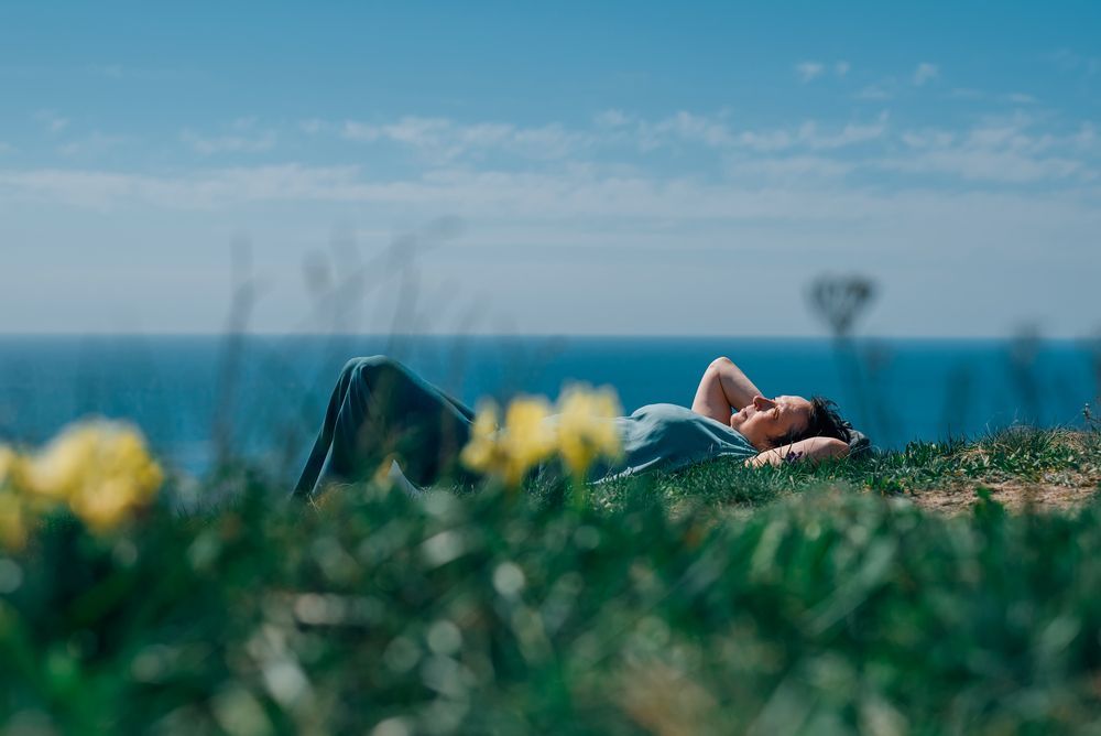 Una mujer yace en una colina cubierta de hierba, mirando el océano bajo un cielo azul.