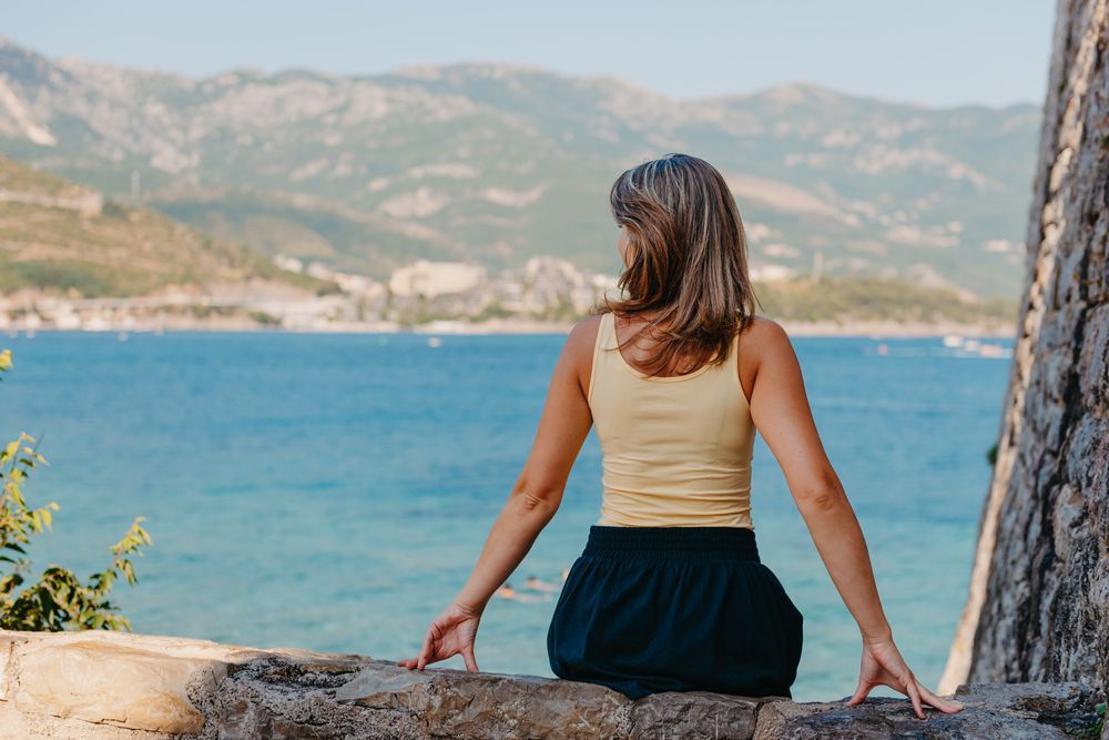 Mujer de cabello gris, con top amarillo y pantalones cortos azules, mira una bahía con montañas desde un muro de piedra.