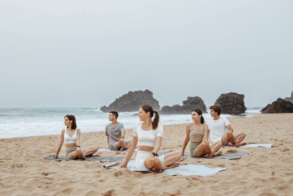 Personas practicando yoga en una playa de arena con rocas al fondo en un día nublado. Están sentadas en torsiones sobre colchonetas.