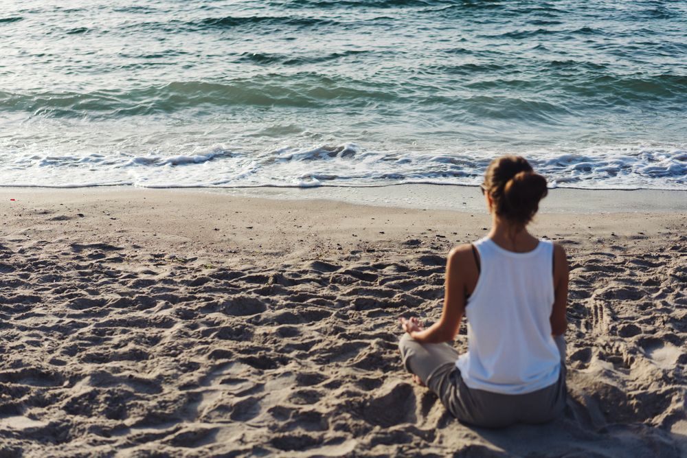 Mujer meditando en una playa de arena frente al mar. Lleva una camiseta blanca sin mangas y pantalones grises.