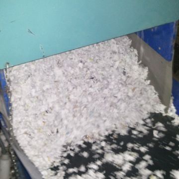 Material blanco triturado amontonado en un contenedor o tolva de plástico azul, con algunos trozos derramados sobre una superficie oscura.