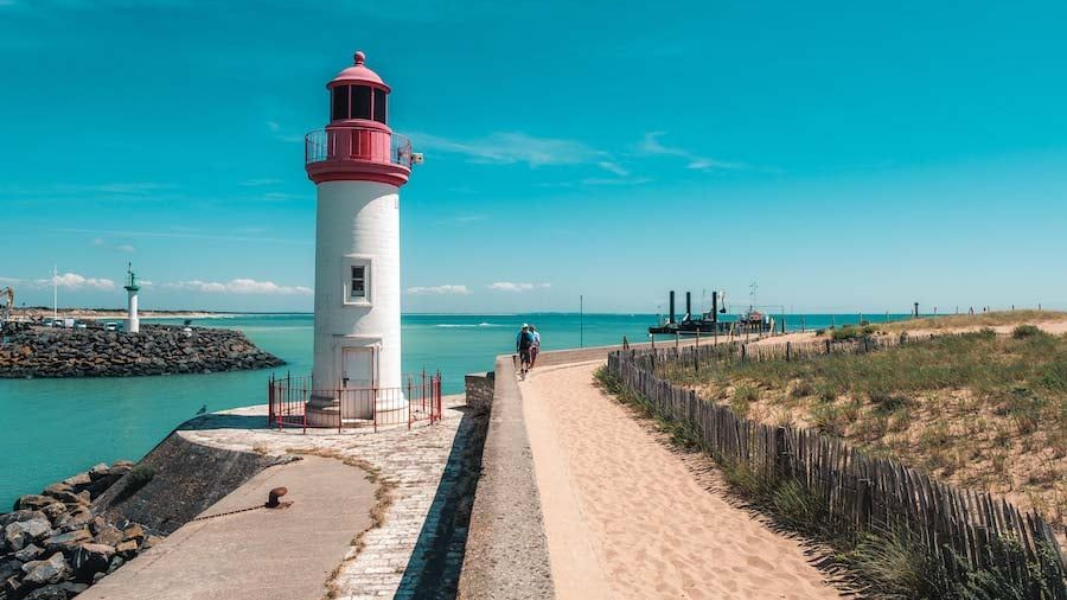 Phare sur une jetée, chemin de sable à droite, mer et ciel bleus.