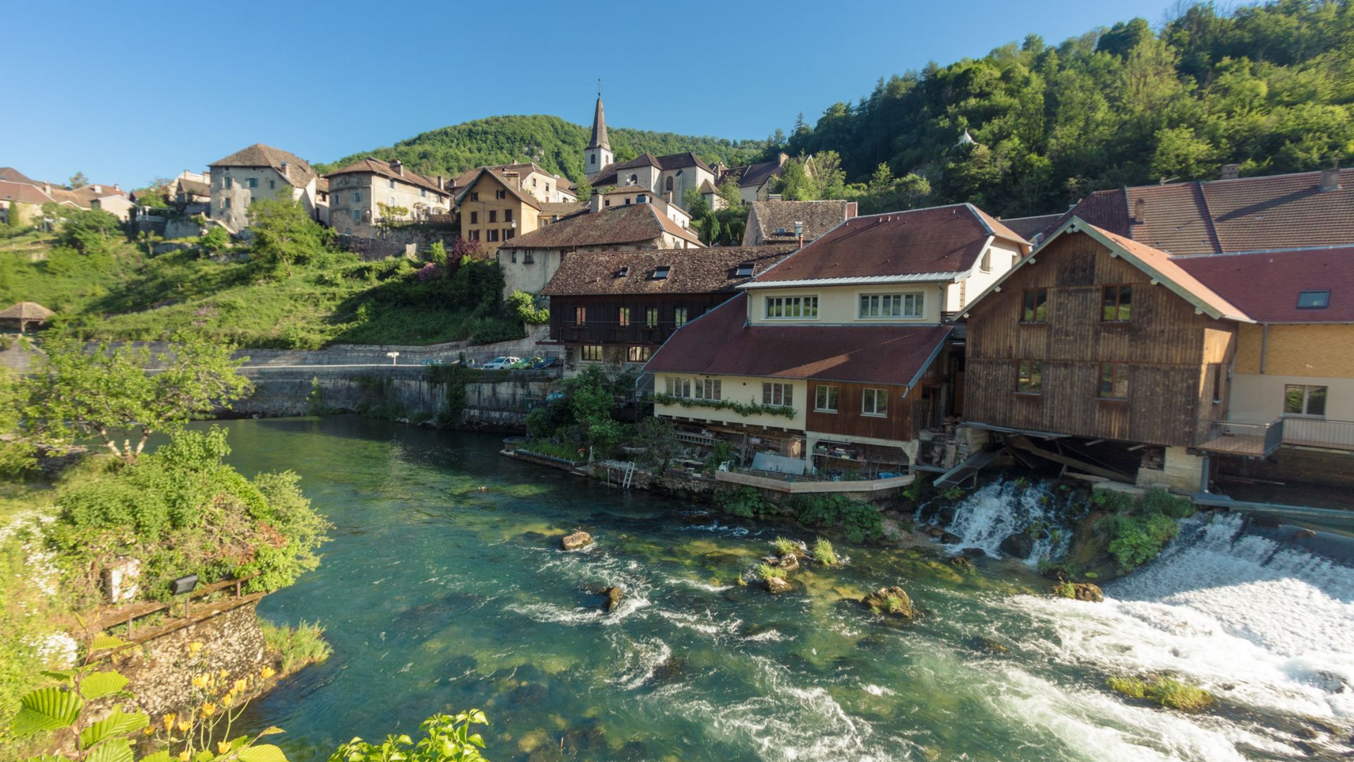 Ville au bord d'une rivière, avec des bâtiments en pierre et en bois et un clocher d'église.