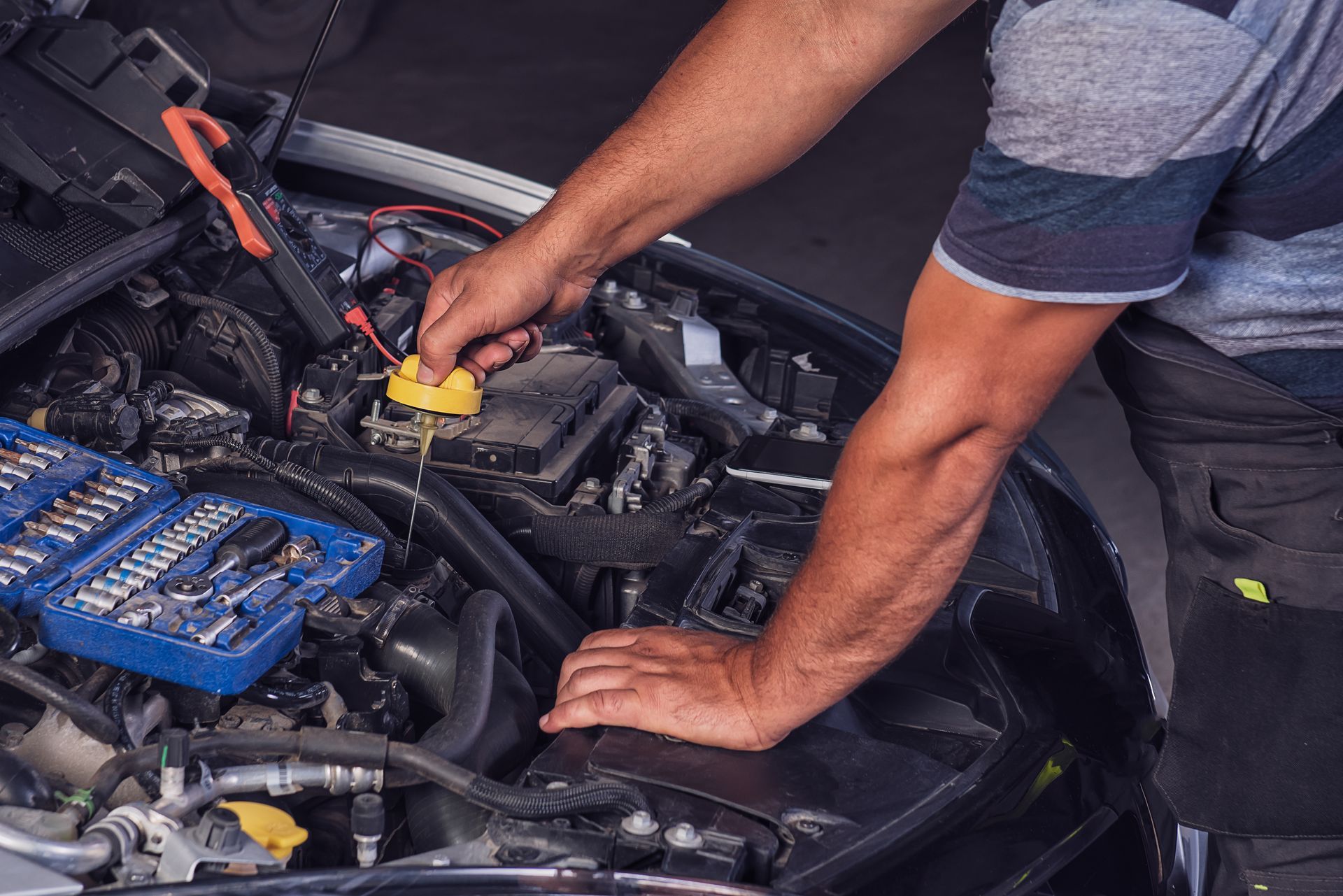 Une personne en tenue de travail se penche dans le compartiment moteur ouvert d'une voiture pour retirer un bouchon de liquide jaune, une trousse à outils se trouve à proximité.