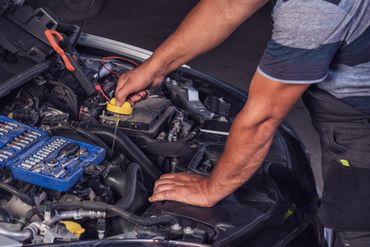 Une personne en tenue de travail se penche dans le compartiment moteur ouvert d'une voiture pour retirer un bouchon de liquide jaune ; une trousse à outils se trouve à proximité.