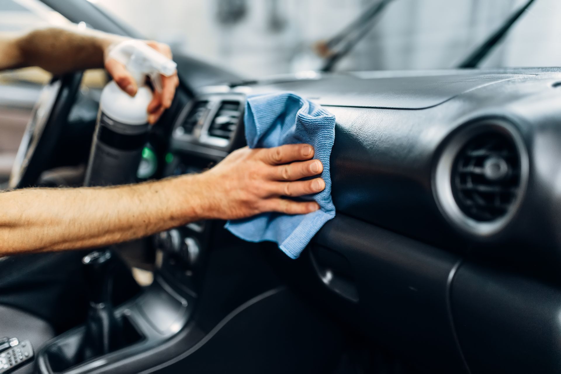 Une personne nettoie le tableau de bord d'une voiture avec un chiffon en microfibre bleu et un flacon pulvérisateur.