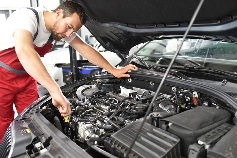 Dans un garage, un mécanicien en uniforme rouge travaille sur le moteur d'une voiture dont le capot est ouvert.