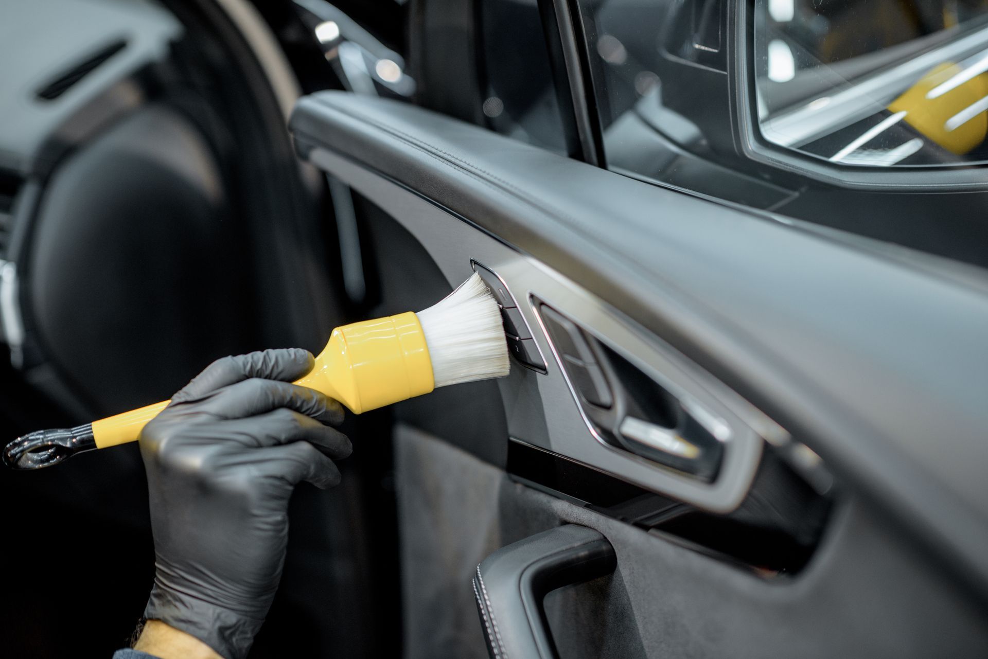 Une personne portant des gants noirs utilise une brosse jaune souple pour nettoyer minutieusement les garnitures intérieures d'une voiture.