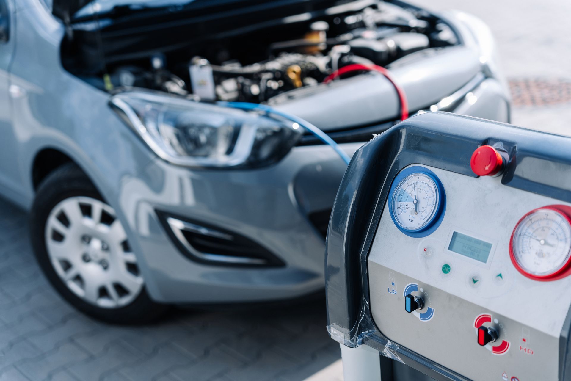 Une machine pour l'entretien de climatisation est connectée à une voiture grise dont le capot est ouvert pour effectuer la maintenance du système de climatisation.