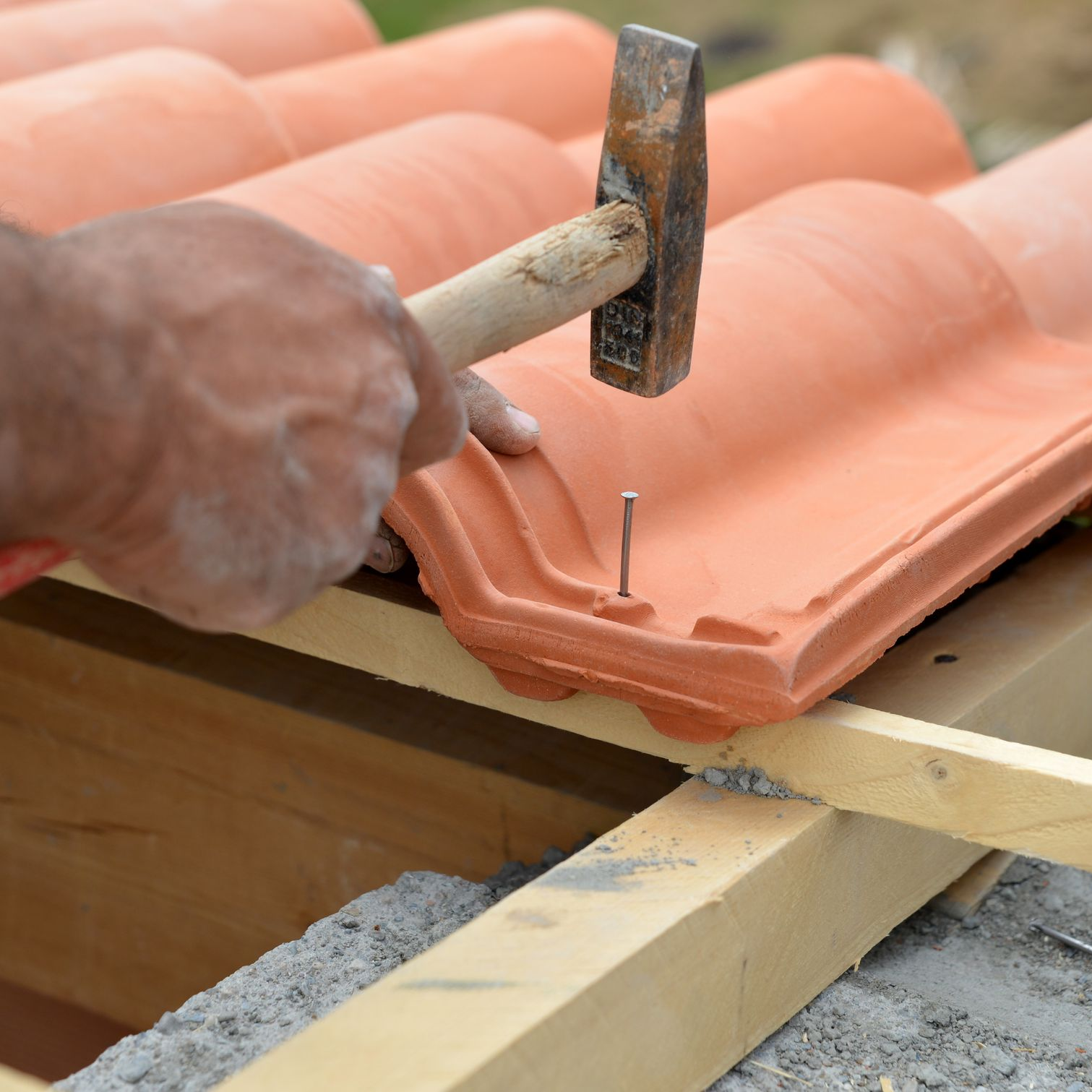Des mains clouent une tuile orange sur des liteaux de bois à l'aide d'un marteau.