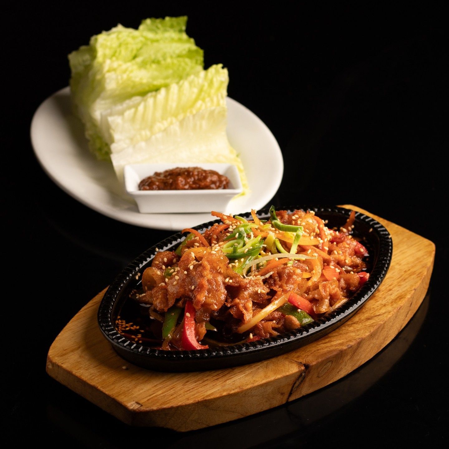 Carne picante salteada en una fuente chisporroteante con verduras cortadas en tiras, junto a hojas de lechuga para hacer wraps, sobre un fondo negro