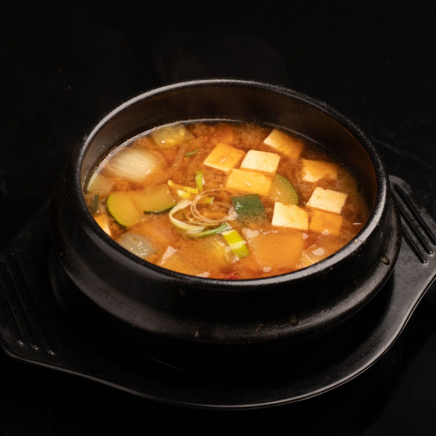 Cuenco negro de sopa de verduras sobre un plato negro, con cubos de tofu y hierbas por encima