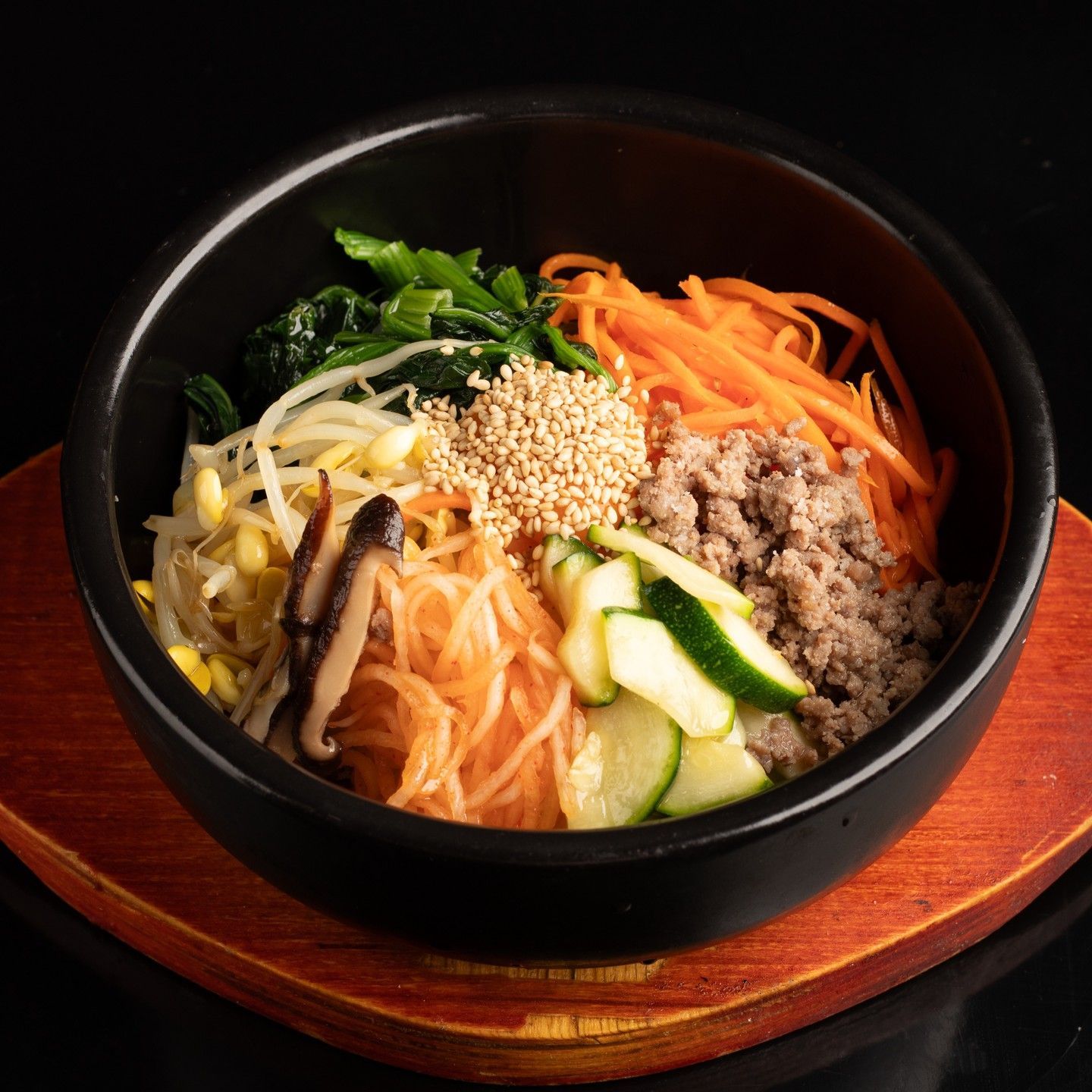 Bibimbap colorido en un cuenco negro sobre una bandeja de madera, cubierto de verduras, carne de res y semillas de sésamo