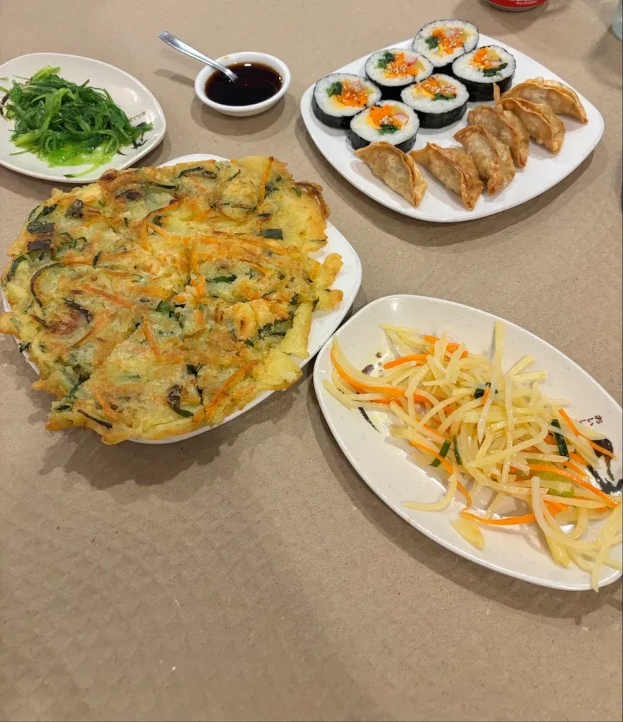 Varios platos coreanos sobre una mesa: tortita de marisco, sushi, empanadillas, ensalada y salsa para mojar.